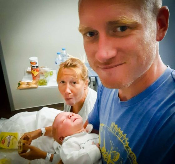 Newborn baby being held by his Dad, while Mum behind eats a sandwich