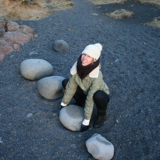 Lady attempting to lift a large rock on a black gravel beach
