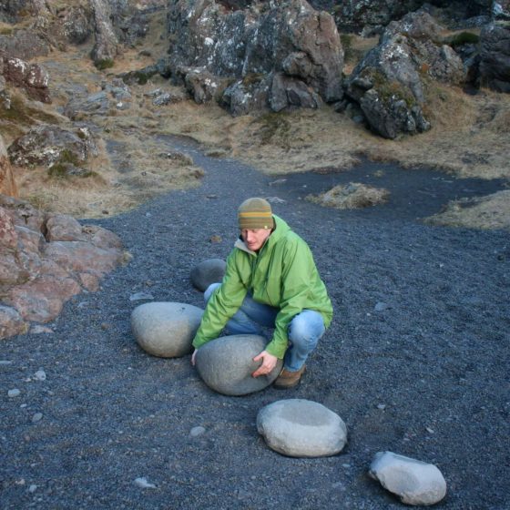 Man attempting to lift a large rock on a black gravel beach