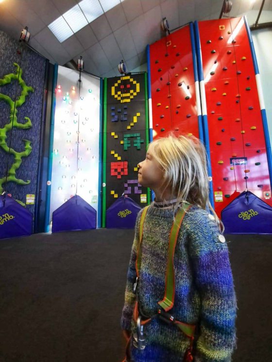 Girl stood in a colourful indoor hall of novelty climbing walls, looking up at the climbs