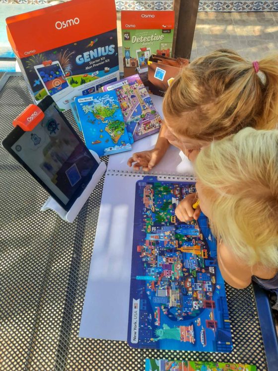 2 girls looking at a cartoon map spread on a table, with an Ipod on a stand behind