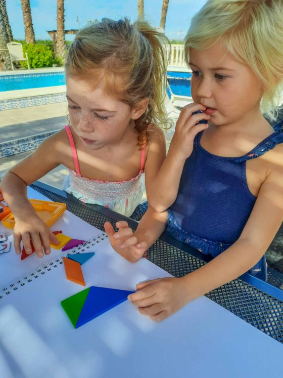 2 girls looking puzzled at an outdoor table, as they try to make a picture out of small wooden shapes