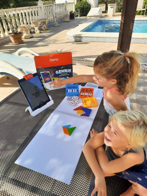 2 girls smiling while sat an outside table, trying to copy a picture on an Ipad screen by assembling small wooden shapes
