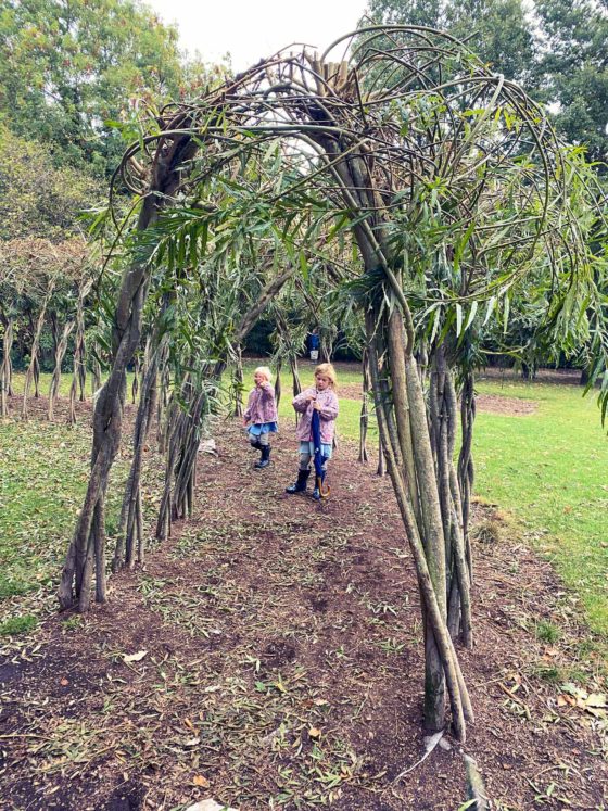 2 girls in wellington boots, stood in an ornamental living Willow tree tunnel in a park