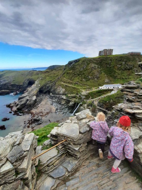 2 young girls wearing raincoats, stood at a castle viewpoint of the coastline below