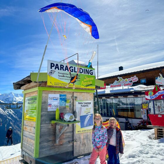 2 girls stood in front of a wooden kiosk in the mountains advertising paragliding