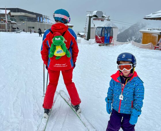 Boy stood next to a ski instructor wearing a dinosaur rucksack
