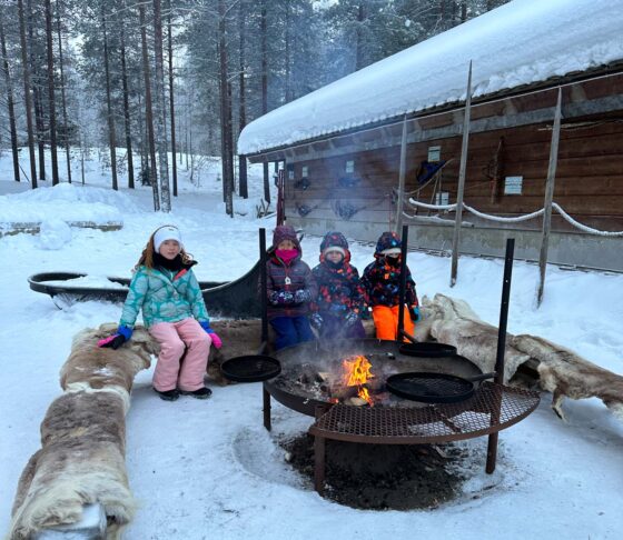4 children wearing thick winter clothes sat around a bonfire in the snow
