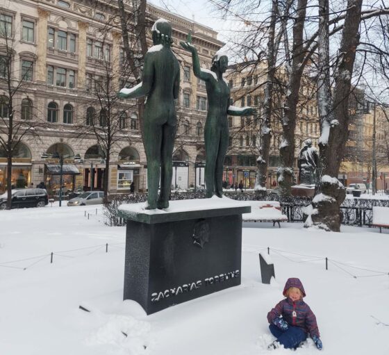 Girl sat in the snow by a stone statue with grand buildings behind