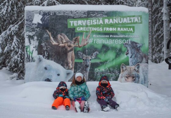 3 small children sat in the snow in front of a sign advertising a winter animal park