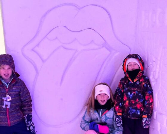 3 children by a snow carving of the Rolling Stones' mouth with tongue out logo