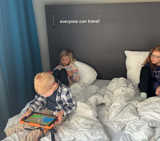 3 children sat on a bed with iPads, with 'everyone can travel' written on the wall behind them